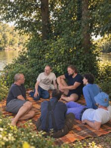 Seven men sitting and talking together on a blanket under a tree beside a river on a summer evening